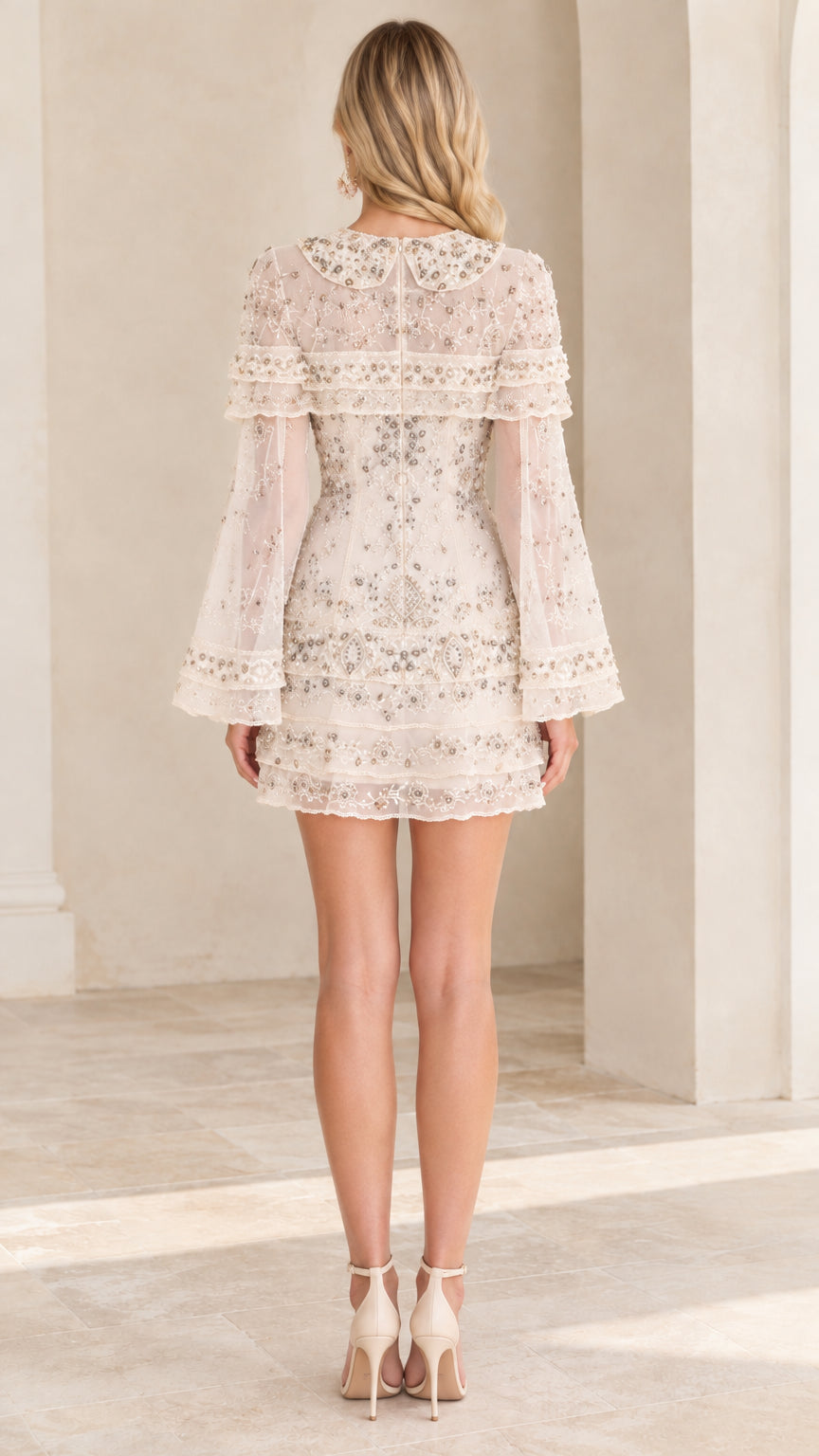 Back view of model wearing cream capelet mini dress with intricate bead embroidery